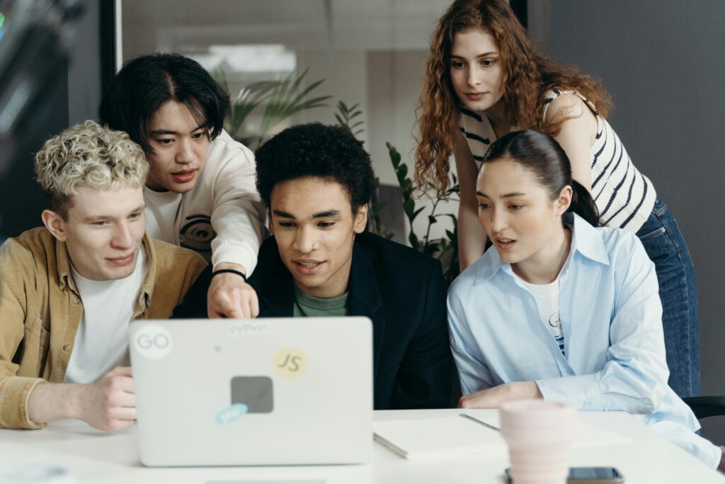 A diverse group of young professionals gathered around a laptop, collaborating on a project related to employee health plans and discussing workplace benefits options.
