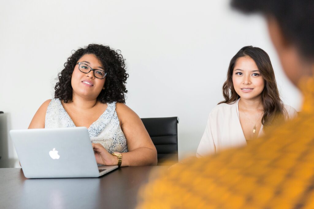 Two women sitting at a conference table with a laptop, actively engaging in a discussion about employee health plans during a workplace consultation.
