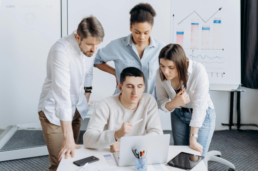 A diverse group of coworkers gathered around a laptop, engaged in analytics with charts in the background, analyzing trends for California insurance plans.