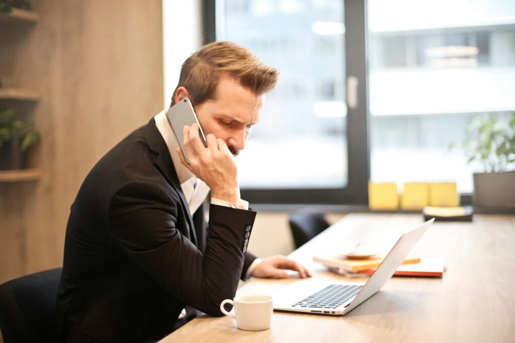 A businessman in a quiet office talks on the phone while reviewing documents on his laptop, researching ACA California enrollment deadlines.