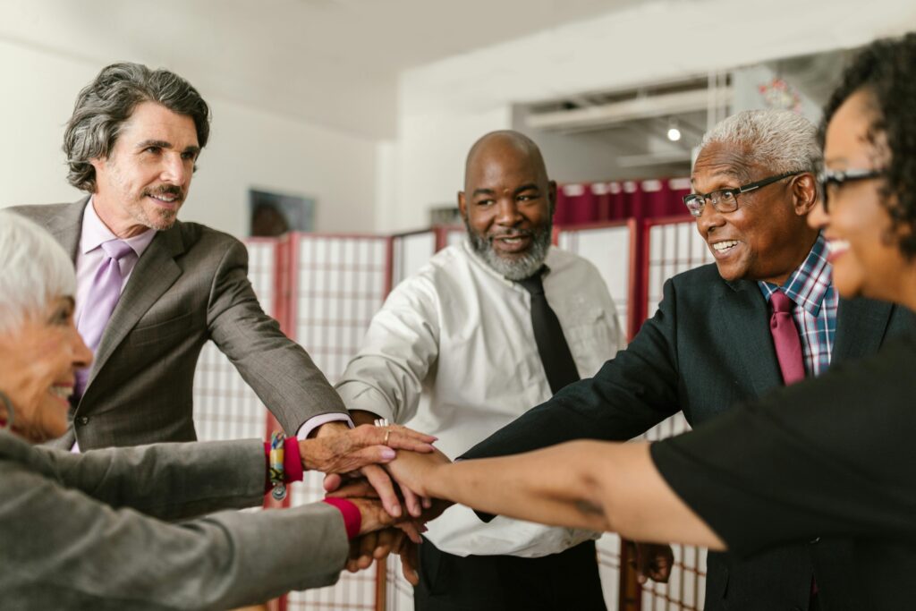 A diverse group of older professionals smiles and stacks hands in unity, symbolizing shared confidence in California insurance plans and Medicare benefits.